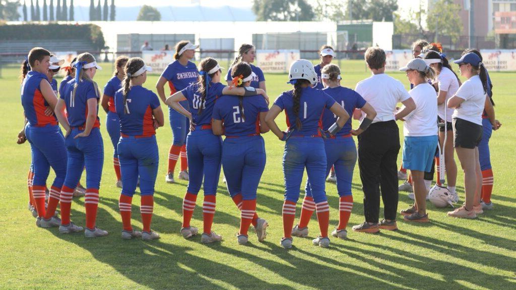 女子垒球队和她们的教练在场地上站成一圈. 球队穿着蓝色的萨基恩队服，配上橙色和白色的袜子.